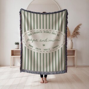 May include: A woven throw blanket featuring vertical sage green and cream stripes with fringed edges. The center displays an oval design with the text "OUR FAVORITE PEOPLE CALL US papa and mimi" and the names "ANNA", "CALEB", "MOLLY", "SAMUEL".