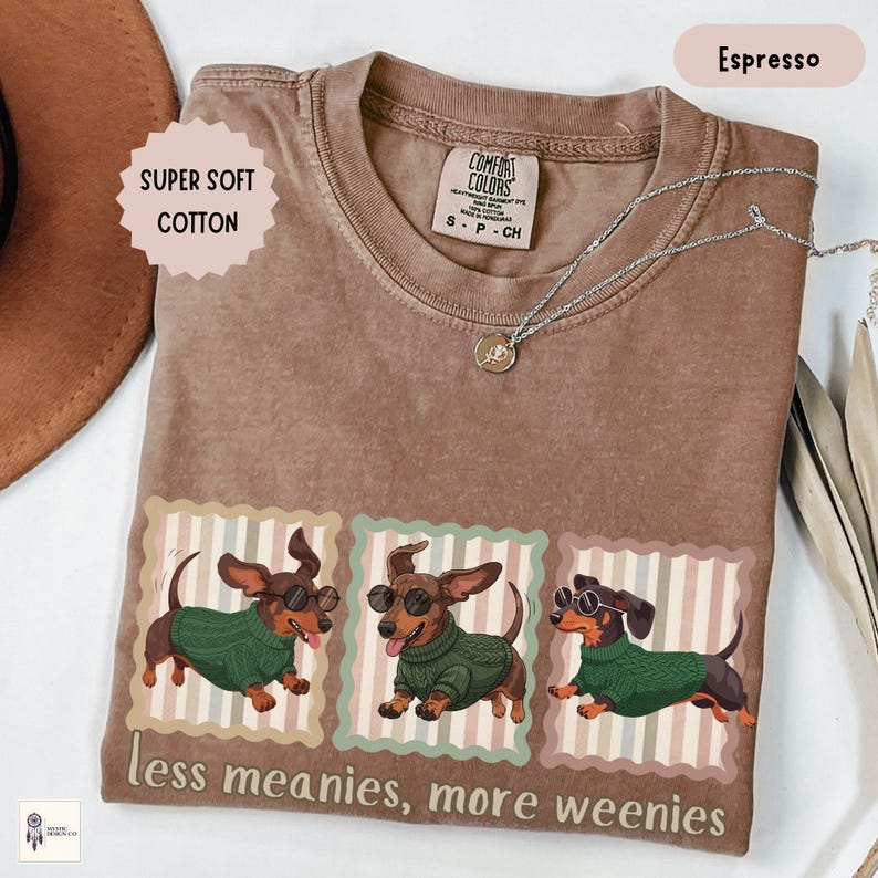 May include: Espresso-colored t-shirt with a graphic of three dachshunds wearing sunglasses and green sweaters. The text "less meanies, more weenies" is printed below the dogs. The shirt also has the text "SUPER SOFT COTTON".