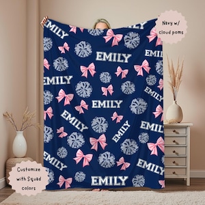 May include: Navy blue blanket with a pattern of pink bows, cloud-like pom-poms, and the name "EMILY" in white. The blanket is customizable with squad colors. Text on the image reads "Navy w/ cloud poms".