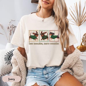 May include: Ivory-colored t-shirt featuring a graphic of three dachshunds wearing sunglasses and green sweaters. The text "less meanies, more weenies" is printed below the dogs. The shirt is a relaxed fit.