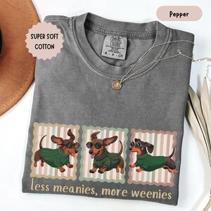 May include: Pepper-colored t-shirt with a graphic of three dachshunds wearing sunglasses and green sweaters. The text "less meanies, more weenies" is printed below the graphic. The shirt also has the text "SUPER SOFT COTTON". A silver necklace is draped across the shirt.