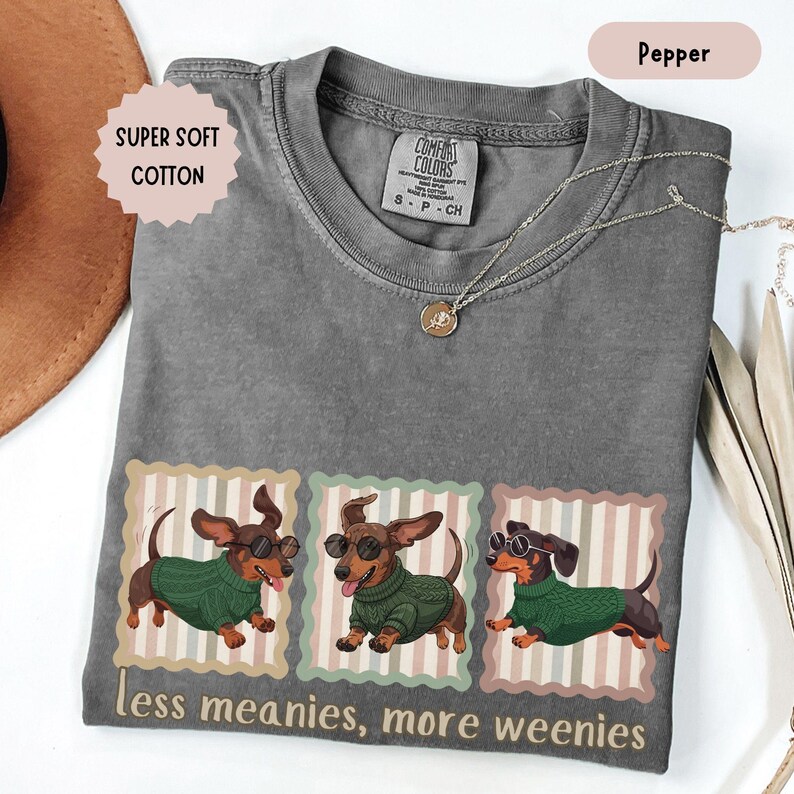 May include: Pepper-colored t-shirt with a graphic of three dachshunds wearing sunglasses and green sweaters. The text "less meanies, more weenies" is printed below the graphic. The shirt also has the text "SUPER SOFT COTTON". A silver necklace is draped across the shirt.