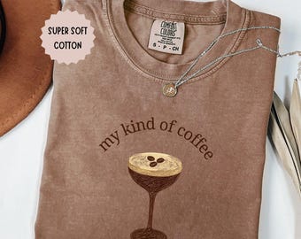 May include: Espresso-colored t-shirt with the phrase "my kind of coffee" and a coffee drink graphic. The shirt has a crew neck and short sleeves. A silver necklace with a pendant is on the shirt. The shirt is labeled "Super Soft Cotton."