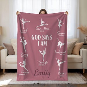 May include: A dusty rose blanket featuring white silhouettes of ballet dancers and inspirational text. The words "God Says I Am" are centered, with phrases like "Loved," "Strong," and "Brave" surrounding the dancers. The name "Emily" is also displayed.