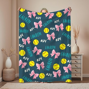 May include: A teal blanket featuring a pattern of yellow softballs, pink bows, the name "LAUREN" in teal script, and the number "24" in white. The blanket is a cozy, personalized item.