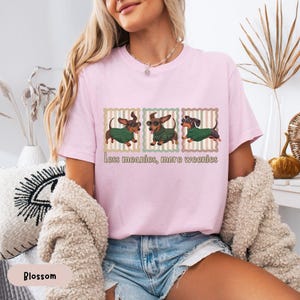 May include: A light pink t-shirt featuring three dachshunds wearing green sweaters and sunglasses. The text "less meanies, more weenies" is printed below the dogs. The shirt is made of a soft material.