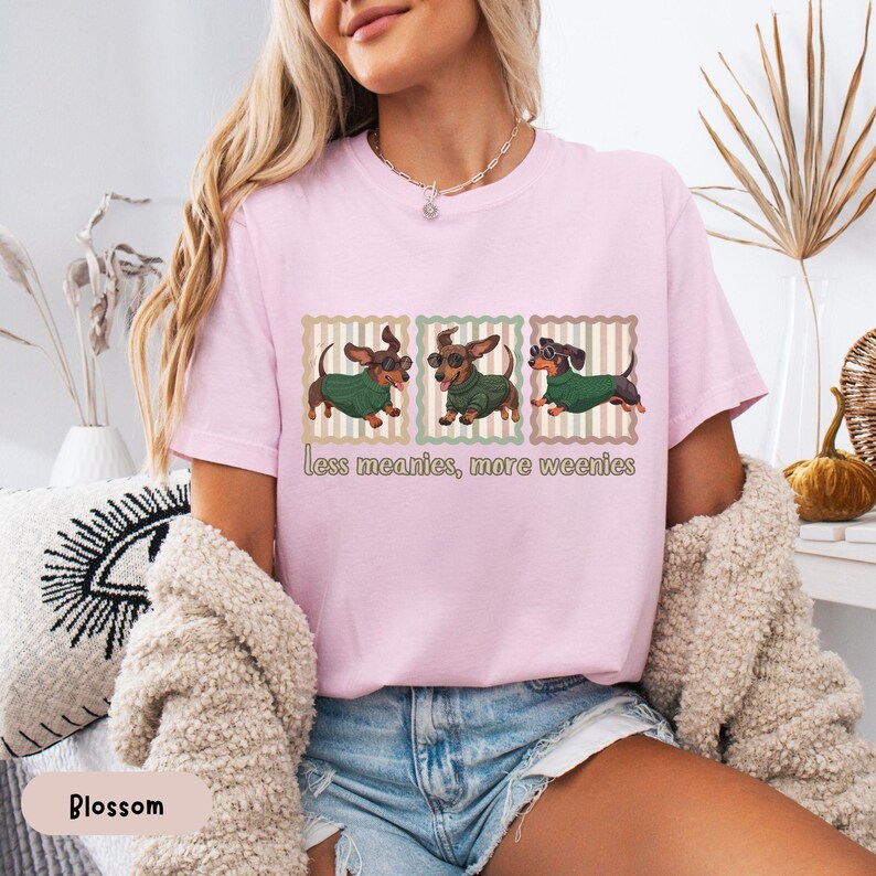 May include: A light pink t-shirt featuring three dachshunds wearing green sweaters and sunglasses. The text "less meanies, more weenies" is printed below the dogs. The shirt is made of a soft material.