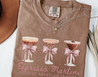 May include: Espresso-colored t-shirt with a graphic of three espresso martinis, each adorned with a pink bow. The text "Espresso Martini BECAUSE ADULTING IS HARD!" is printed below the graphic. A silver necklace is draped across the shirt.