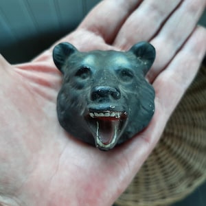 May include: A black bear head figurine with an open mouth, revealing teeth and a red interior. The bear's face is detailed with visible fur texture and is held in a hand.