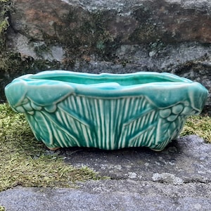 May include: A turquoise ceramic planter with a rectangular shape and a detailed leaf design. The planter has a textured surface with vertical lines and a scalloped rim. The planter is sitting on a stone surface.