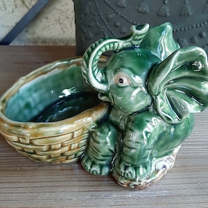 May include: A green ceramic elephant planter with a basket-like base. The elephant has large ears and a trunk curled up. The planter is perfect for small plants or succulents.