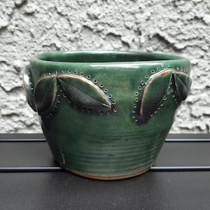 May include: A handcrafted, dark green ceramic bowl with leaf-shaped accents. The bowl features a textured surface and decorative perforations along the rim. The leaves are a darker shade of green with brown detailing. The bowl is sitting on a black surface.