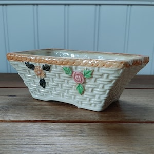 May include: A rectangular ceramic basket planter with a woven texture. The planter is off-white with a light brown rim and floral accents. The flowers are pink, orange, and green. The planter is sitting on a wooden surface.