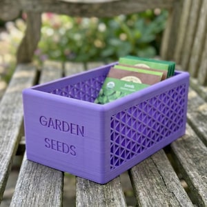 May include: A purple rectangular container with the words "GARDEN SEEDS" embossed on the side. The container has a lattice design and holds several seed packets. The container is sitting on a wooden bench.