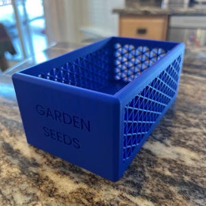 May include: A vibrant blue rectangular container with the words "GARDEN SEEDS" embossed on the front. The sides feature a geometric lattice design. This container is likely used for storing garden seeds.