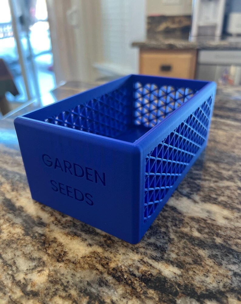 May include: A vibrant blue rectangular container with the words "GARDEN SEEDS" embossed on the front. The sides feature a geometric lattice design. This container is likely used for storing garden seeds.