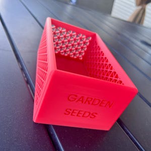May include: A bright red, rectangular container with the words "GARDEN SEEDS" embossed on the side. The container has a lattice design on the sides and a geometric pattern on the top interior. The container is sitting on a dark surface.