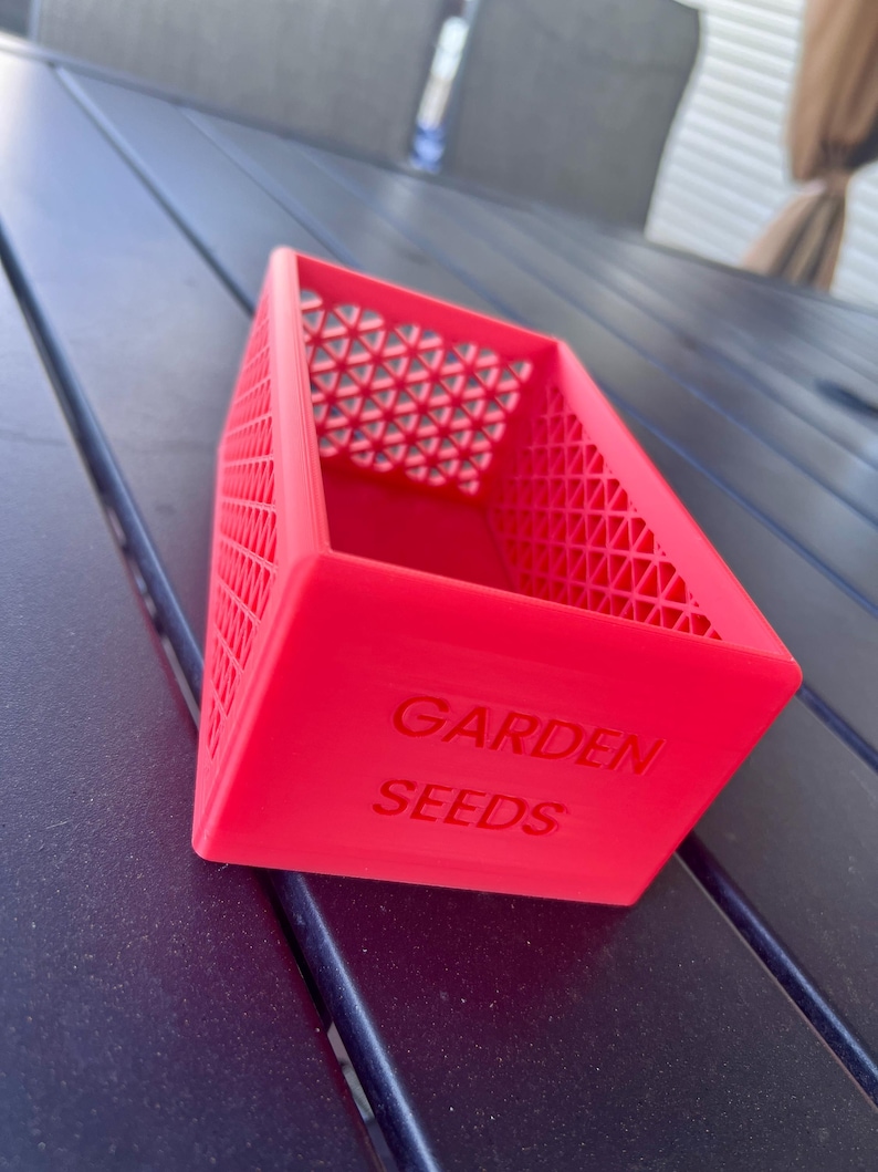 May include: A bright red, rectangular container with the words "GARDEN SEEDS" embossed on the side. The container has a lattice design on the sides and a geometric pattern on the top interior. The container is sitting on a dark surface.