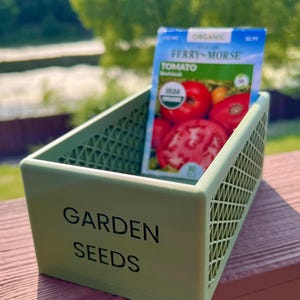 May include: A light green plastic container labeled "GARDEN SEEDS" holds a packet of Ferry-Morse organic tomato seeds. The packet features images of ripe tomatoes. The container has a lattice design.