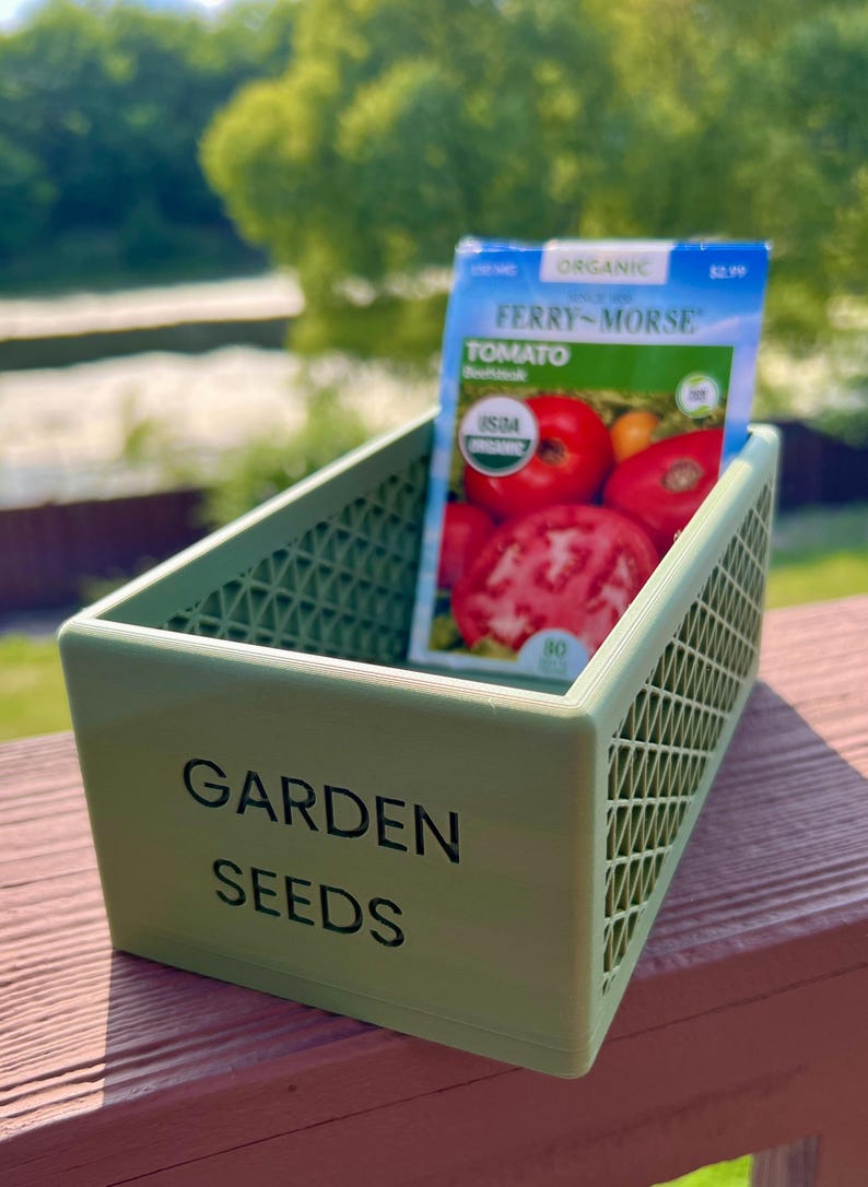 May include: A light green plastic container labeled "GARDEN SEEDS" holds a packet of Ferry-Morse organic tomato seeds. The packet features images of ripe tomatoes. The container has a lattice design.