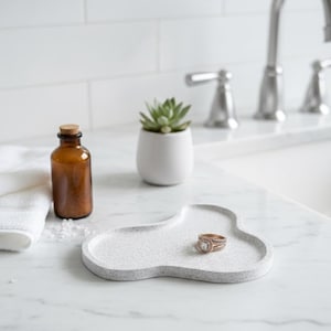 May include: A speckled gray ceramic tray with a gold ring sits on a white countertop. A small brown bottle with a cork stopper and a white potted succulent are also on the counter. A white towel is visible.