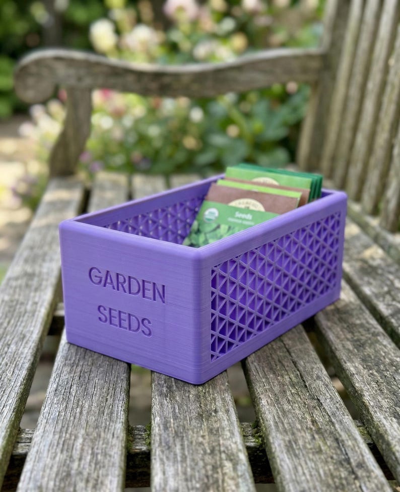 May include: A purple rectangular container with the words "GARDEN SEEDS" embossed on the side. The container has a lattice design and holds several seed packets. The container is sitting on a wooden bench.