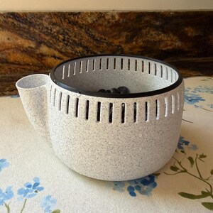 May include: A round, speckled white self-watering planter with a black rim and a side water reservoir spout. The planter features vertical ventilation slits around the upper perimeter and contains dark soil or stones visible inside the basin.