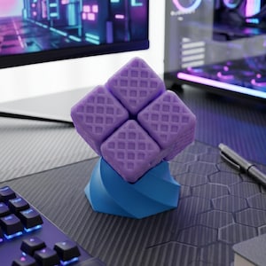 May include: A purple cube-shaped desk toy with a waffle pattern, atop a blue twisted base. The toy sits on a dark gray desk with a keyboard, pen, and a computer monitor in the background.