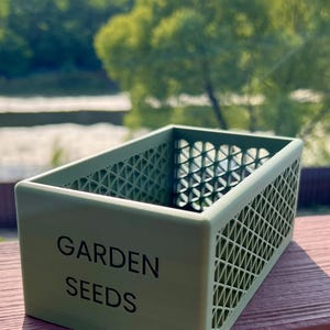 May include: A light green rectangular container with the words "GARDEN SEEDS" printed on the front. The container has a lattice design on the sides and is sitting on a wooden surface. The background features a blurred view of trees and a body of water.