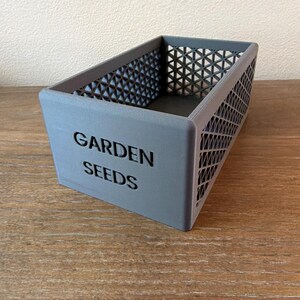 May include: A gray rectangular container with the words "GARDEN SEEDS" printed on the front. The sides have a geometric pattern. The container is sitting on a wooden surface.