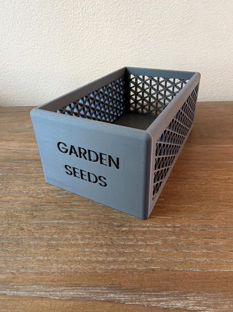 May include: A gray rectangular container with the words "GARDEN SEEDS" printed on the front. The sides have a geometric pattern. The container is sitting on a wooden surface.
