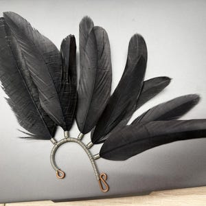 May include: An ear cuff with dark grey feathers attached to a silver wire frame. The feathers are arranged in a fan shape, with copper-coloured accents at the base. The design is suitable for a special occasion.