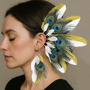 May include: A decorative ear cuff and matching earring set featuring peacock feathers. The design incorporates white, gold glitter, and vibrant peacock eye details in blue, green, and black. The ear cuff wraps around the ear, while the earring dangles.