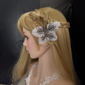 May include: A butterfly-shaped hair clip made of white and speckled feathers. The clip is attached to a mannequin with blonde hair styled in a braid. The butterfly has a silver center and black antennae.