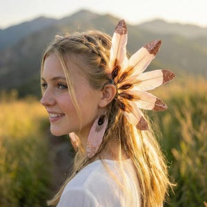 May include: A hair accessory featuring a cluster of light-coloured feathers with glittered tips. The feathers are arranged in a fan shape, attached to a hair clip. The accessory is worn on the ear, adding a touch of bohemian flair.