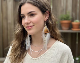 May include: Feather earrings with a gold-colored hook. One earring features a triangular section of brown and black feathers, while the other is white. Both earrings have long, white and tan feathers.