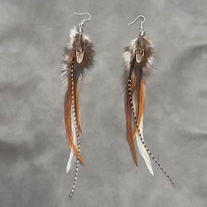 May include: A pair of feather earrings with silver-tone hooks. The earrings feature a cluster of brown and white feathers at the top, with long, flowing feathers in shades of brown, orange, and white. The earrings have a bohemian style.