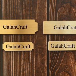 May include: Four brass-coloured nameplates with the text "GalahCraft" in a black, sans-serif font. The nameplates are rectangular, with two having decorative edges. The background is a dark brown wooden surface.