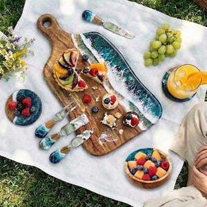 May include: A picnic scene with a wooden cutting board, serving knives, and a decorative serving tray with a blue and white wave design. The board holds fruit tarts and berries. A glass of orange juice and a bowl of fruit are also present.