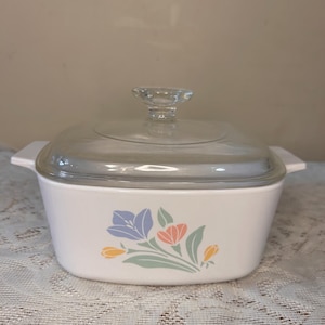 Op de afbeelding: Witte vierkante ovenschaal met een helder glazen deksel en handvat. De schaal heeft een bloemenontwerp in blauw, oranje, geel en groen. De schaal heeft aan weerszijden twee kleine handvatten.