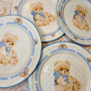 May include: Set of four cream-colored plates with a teddy bear design. Each plate features a large teddy bear with a blue bow, surrounded by a blue border with small teddy bear and floral accents. A charming addition to any kitchen.