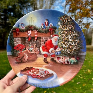 May include: A decorative plate featuring a Christmas scene with Santa Claus, a decorated tree, and a fireplace. The plate has a blue border and depicts a festive interior with gifts and a cat. A tray with cookies and a mug is in the foreground.