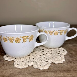 Può includere: Due tazze da tè in ceramica bianca con motivi floreali dorati sul bordo. Le tazze hanno grandi manici e poggiano su un centrino all'uncinetto color crema. Lo sfondo è beige neutro.