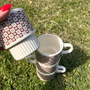 May include: A set of four stackable ceramic mugs featuring a cream-colored base with a repeating brown grid pattern containing small white floral motifs. The mugs have a smooth interior and a ribbed texture on the lower exterior section.