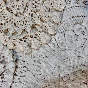 May include: Close-up of off-white crocheted doilies and lace fabric. The doilies have a circular design with detailed patterns. The lace fabric has a delicate, open weave. The textures and patterns create a vintage, handmade aesthetic.