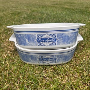 May include: Two white ceramic baking dishes stacked vertically, featuring blue marble-patterned bands and a central rectangular graphic with a blue checkerboard border. The text reads "The Art of Bon Appetit Fine Cuisine" in a script font.