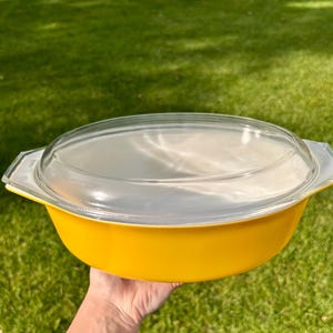May include: A bright yellow oval casserole dish with a clear glass lid. The dish features integrated handles on both sides and a smooth, solid-colored exterior. The lid is transparent, domed, and fits securely over the top of the yellow base.
