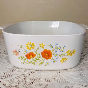 May include: White rectangular casserole dish with a floral design. The dish features orange poppies, yellow daisies, and blue forget-me-nots. The dish has two handles and is set on a white lace tablecloth.