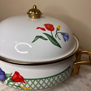 Puede incluir: Olla de cocina de esmalte blanco con tapa, con un diseño floral de tulipanes rojos, flores azules y hojas verdes. La olla tiene asas doradas y un pomo dorado en la tapa. Un patrón de celosía verde decora la base.