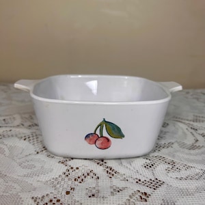 May include: White square casserole dish with two handles. The dish features a painted design of two red cherries with green leaves. The dish is sitting on a white lace tablecloth.